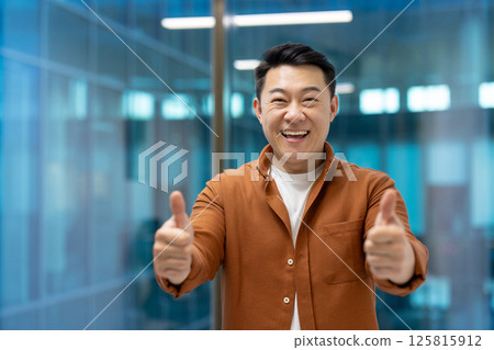 Smiling Asian businessman in office showing thumbs up expressing positivity and approval. Casual attire and cheerful expression create joyful, motivational ambiance in corporate environment. 125815912