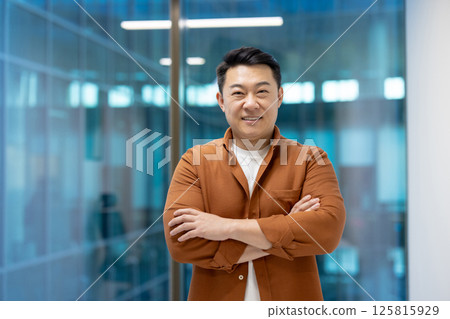 Asian businessman confidently stands with arms crossed in modern office. Casual attire highlights approachable leadership. Expression conveys professionalism, confidence, and executive presence. 125815929