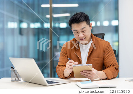 An asian businessman sits at his desk, making notes in a notebook while working on a laptop in a modern office setting. 125815950