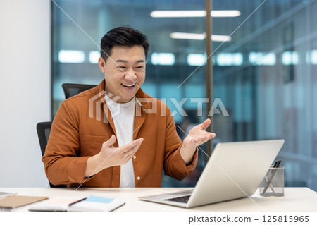 A happy asian man is sitting in an office, smiling and gesturing during a video call on his laptop. Modern office setting. 125815965