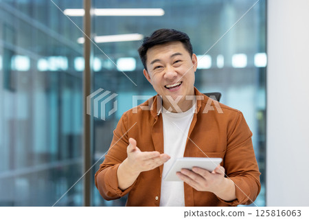 Happy asian man in a bright office setting holding a tablet and gesturing with his hand, smiling. 125816063