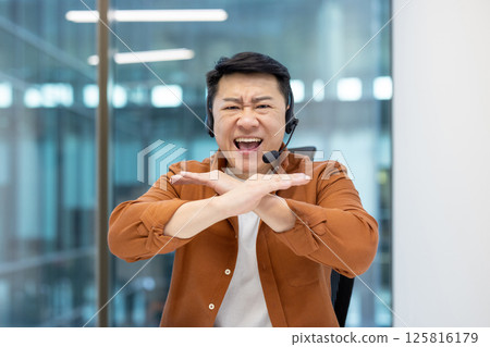 Webcam view, Asian man with headset quarreling angry shouting at camera. Dissatisfied support worker can't understand client. Man at workplace inside office. 125816179