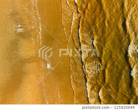 Aerial view of ocean waves gently rolling onto a sandy beach, creating natural patterns and textures in the shoreline. The image captures the contrast between water and land from above. 125820844