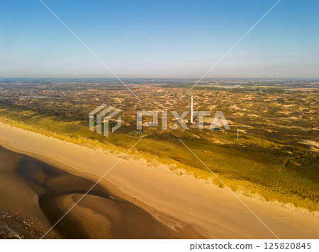 A stunning aerial view of a long sandy beach stretching along the coast bordered by dunes and greenery with a clear blue sky and calm sea creating peaceful natural atmosphere. Tall communication tower 125820845