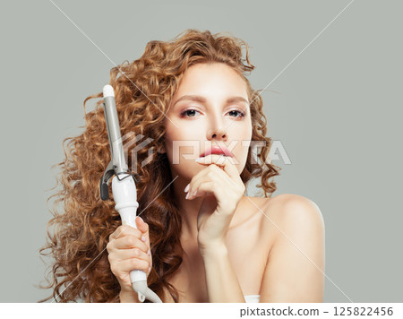 Sensual brown haired woman with shiny long curly hairstyle showing curling iron. Sensual brown haired woman with shiny long curly hairstyle showing curling iron. 125822456