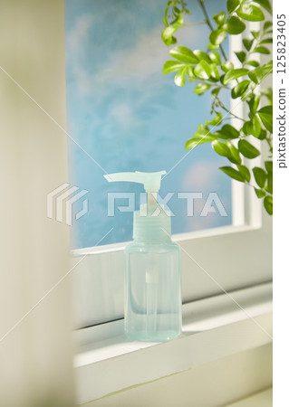 A blue cosmetic bottle with a pump stands on a white window shelf, next to leafy green plants. Inside the bottle is a clear liquid essence. Outside the glass door is a view of the clear blue sky. 125823405