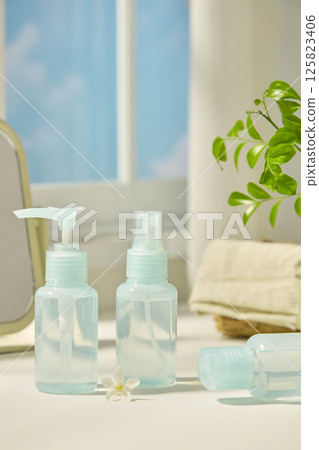 Composition of cosmetic bottle samples. A bottle lies on table, a daisy placed in the center for decoration. The tree branches with many green leaves, a wicker basket and a mirror blurred in behind. Composition of cosmetic bottle samples. A bottle lies on table, a daisy placed in the center for decoration. The tree branches with many green leaves, a wicker basket and a mirror blurred in behind. 125823406