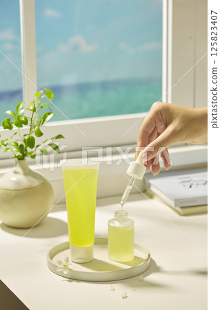 A tube filled with yellow liquid extracted from lemon. Hand holding the pump of a dropper cosmetic bottle, drops of essence are flowing into the glass bottle. Scene of a table next to the window. 125823407