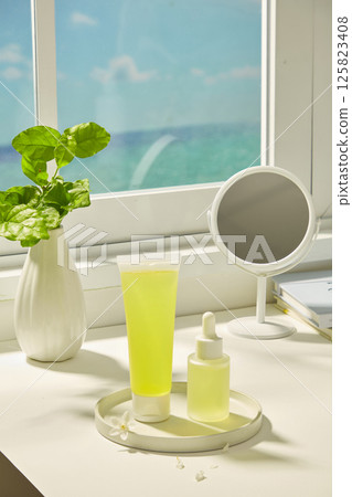 Vertical photo of tubes and dropper bottles placed on a stone plate. Slender vase with green leaves. Table with mirror and books placed next to the window with a view of the blue sky outside. 125823408