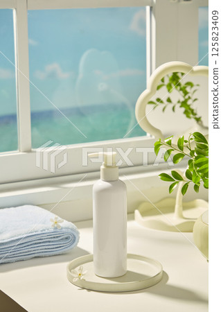Tall cosmetic bottles with pump dispensers on round stone pedestals, displayed on white table. Neatly folded blue towels, white daisies scattered around. White windows with sea views outside. 125823409