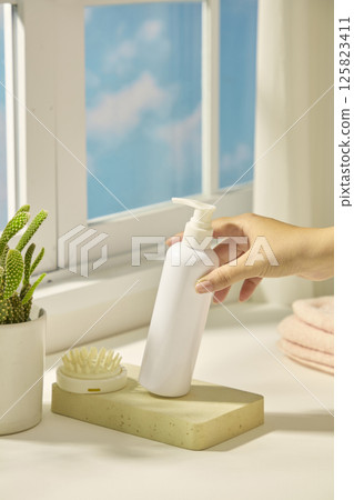 A slender hand tilts a bottle of cosmetics. A hand comb sits on a rectangular stone slab, next to a cactus pot. Light from the window shines in the scene, shadows of objects fall on the white table. A slender hand tilts a bottle of cosmetics. A hand comb sits on a rectangular stone slab, next to a cactus pot. Light from the window shines in the scene, shadows of objects fall on the white table. 125823411
