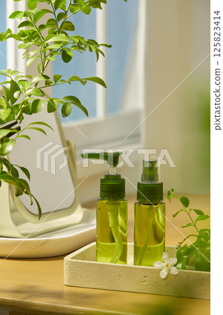 Two transparent green cosmetic bottles in travel sample form, placed on a polished stone tray. A table mirror placed on a tray behind, along with a lush houseplant. A few daisies decorated around. Two transparent green cosmetic bottles in travel sample form, placed on a polished stone tray. A table mirror placed on a tray behind, along with a lush houseplant. A few daisies decorated around. 125823414