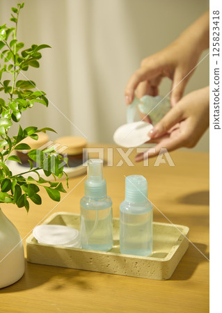 Hands pumping micellar water onto cotton pads. Small blue cosmetic bottles filled with essence, displayed on rectangular trays. Porcelain vases holding leafy green plants. Lifestyle concept.  125823418