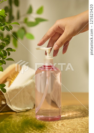 Fingers of hand touch the pump head of a pink cosmetic bottle, filled with liquid and placed on a straw mat, next to a wicker basket containing cosmetic tubes, a wooden comb and coarse cloth. Fingers of hand touch the pump head of a pink cosmetic bottle, filled with liquid and placed on a straw mat, next to a wicker basket containing cosmetic tubes, a wooden comb and coarse cloth. 125823420