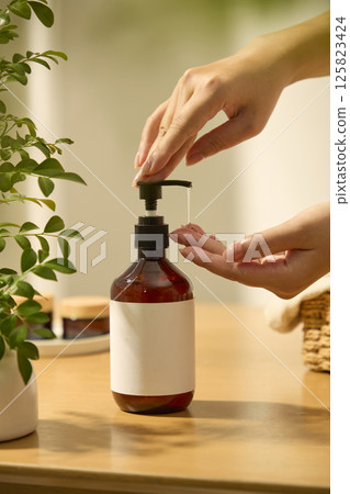 A hand gently presses the pump nozzle of the bottle, the essence flows out from the amber bottle, another hand is catching the essence. Frontal shot of an unlabeled bottle, used for advertising. 125823424