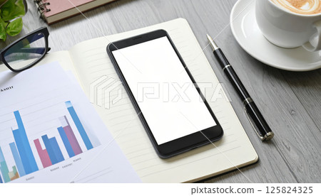 blank smartphone screen is displayed on open journal, surrounded by pen, documents, coffee cup, and pair of glasses, creating productive workspace atmosphere 125824325