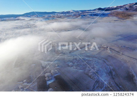Aerial view of snow-covered landscape with fog rolling over fields and hills in winter season 125824374