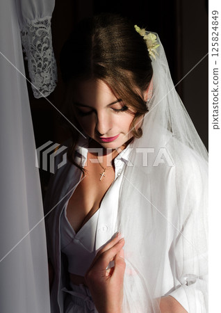 Bride in white robe, adjusting her veil. Sunlight streams in, casting shadows. A moment of quiet contemplation before the ceremony. Bride in white robe, adjusting her veil. Sunlight streams in, casting shadows. A moment of quiet contemplation before the ceremony. 125824849
