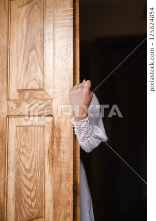 A hand emerges from behind a wooden door, a glimpse of a white dress visible. A hand emerges from behind a wooden door, a glimpse of a white dress visible. 125824854