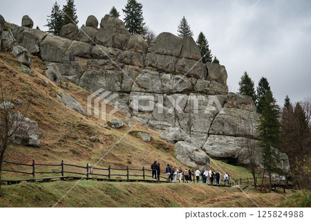 Hikers explore a unique rock formation on a hillside, surrounded by lush greenery and tall trees. 125824988