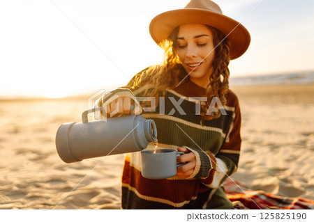 Young woman sits at picnic on the beach drinks a hot drink from a thermos. Travel, weekend, relax Young woman sits at picnic on the beach drinks a hot drink from a thermos. Travel, weekend, relax 125825190