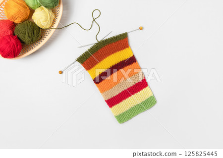 Red, green, orange and beige knitting balls in basket with knitting needles and knitted scarf on gray background. Set for the hobby of knitting warm clothes or toys. Concept needlework 125825445