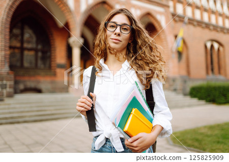 Woman student holds several colorful notebooks, standing outside university. University Student. 125825909