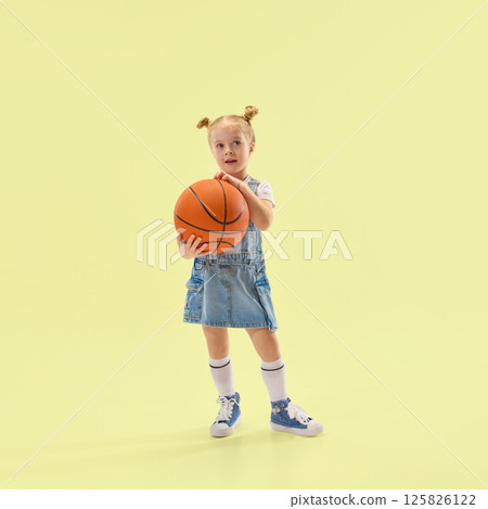 Young girl holds basketball, wearing denim dress and white socks, looking curious and calm, posing against yellow backdrop 125826122