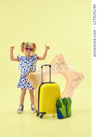 Cheerfully girl raising hands in excitement, standing near suitcase and inflatable float, expressing happiness against yellow background. 125826146