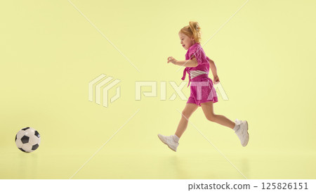 Joyful child girl in pink shorts and shirts running towards soccer ball, playing against yellow background. Joyful child girl in pink shorts and shirts running towards soccer ball, playing against yellow background. 125826151