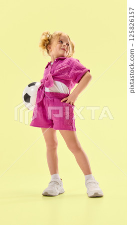 Full-length low angle view of happy smiling child girl wearing pink summer outfit, posing with soccer ball against light yellow background. Full-length low angle view of happy smiling child girl wearing pink summer outfit, posing with soccer ball against light yellow background. 125826157