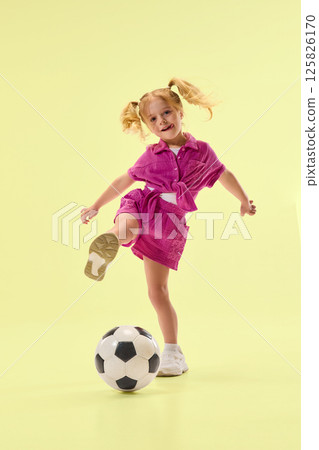 Smiling girl in pink outfit joyfully playing with soccer ball on pastel yellow background. Active lifestyle of kid. 125826170
