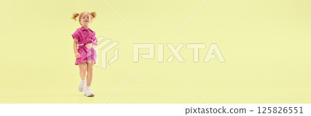 Low angle view of happy child girl with ponytail hairstyle, walking in pink outfit, smiling playfully against yellow background. Low angle view of happy child girl with ponytail hairstyle, walking in pink outfit, smiling playfully against yellow background. 125826551