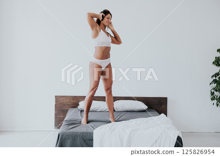 Brunette in lingerie listening to music in headphones and dancing in bedroom Brunette in lingerie listening to music in headphones and dancing in bedroom 125826954