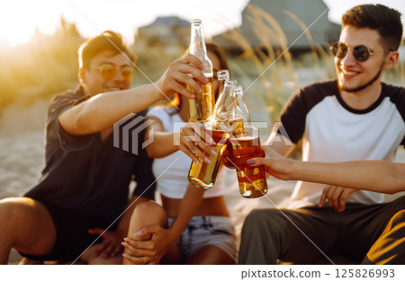 Young people sitting together at beach, drinking beer. Group of friends cheers with beers at the beach. Young people sitting together at beach, drinking beer. Group of friends cheers with beers at the beach. 125826993