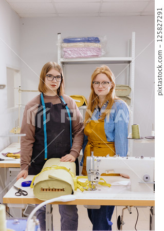 Two female seamstresses in workshop at factory are standing at a table with sewing machines.  125827291