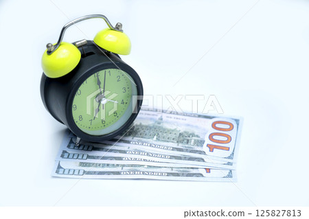 Green alarm clock and a pile of 100 dollar banknotes on a white background. Concept of time is money, saving or investment, and tax Green alarm clock and a pile of 100 dollar banknotes on a white background. Concept of time is money, saving or investment, and tax 125827813