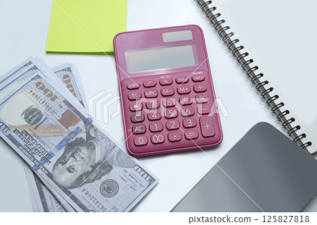 Pink calculator and a pile of 100 dollar banknotes with a notebook, notes, and a cellphone with a white background. Mockup or Copy space. Concept of wealth, saving or investment, and tax 125827818