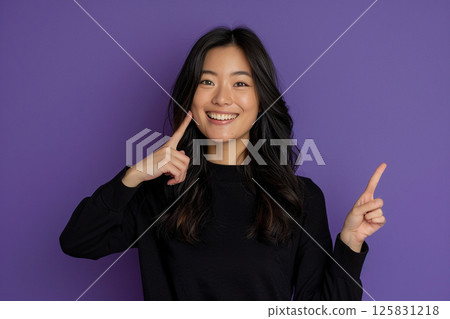 happy young woman pointing up, isolated on a purple background, in side view. Mockup for text and design. Asian girl wearing a white t-shirt, smiling and posing in a studio. 125831218