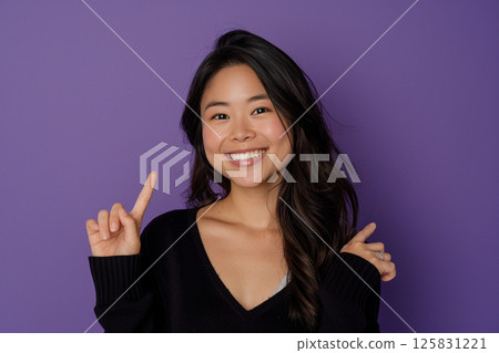 happy young woman pointing up, isolated on a purple background, in side view. Mockup for text and design. Asian girl wearing a white t-shirt, smiling and posing in a studio. 125831221