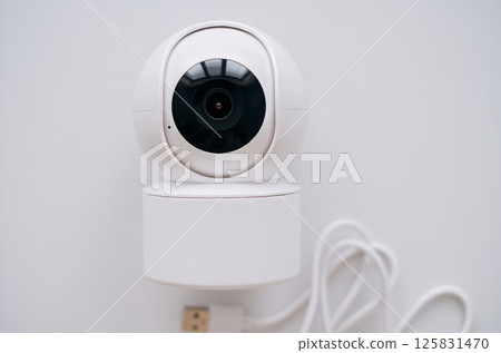 Close-up image of a modern white smart security camera mounted on a wall with a USB cable attached 125831470