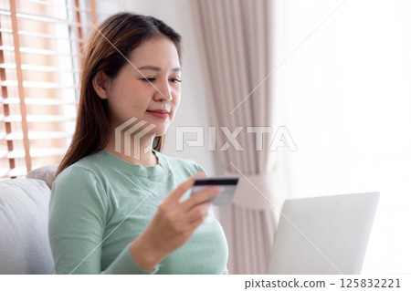 Young asian woman sitting on sofa using laptop computer shopping online with credit card. 125832221