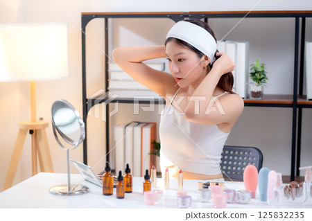 Young asian woman wearing headband and tying hair while preparing skincare looking mirror in room. Young asian woman wearing headband and tying hair while preparing skincare looking mirror in room. 125832235