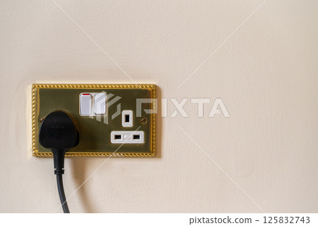UK style double electrical socket with plug inserted. Gold socket with white details, black plug and magnolia wall. One switch in on position. No branding. Copy space to right. UK style double electrical socket with plug inserted. Gold socket with white details, black plug and magnolia wall. One switch in on position. No branding. Copy space to right. 125832743