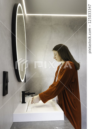 Young woman in a red robe rinsing her hands under running water in a modern bathroom with soft lighting and a glowing round mirror. Young woman in a red robe rinsing her hands under running water in a modern bathroom with soft lighting and a glowing round mirror. 125833347