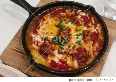 close up of baked meatball in tomato sauce with cheese toppings in a pan on wooden insulating display board 125834047