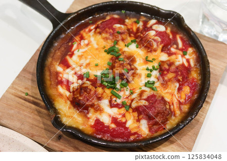 close up of baked meatball in tomato sauce with cheese toppings in a pan on wooden insulating display board 125834048