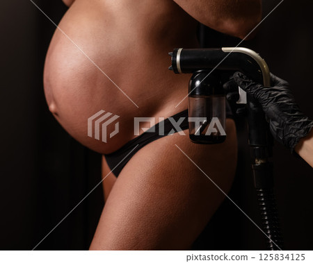 Close-up of the belly of a pregnant woman undergoing an instant tanning procedure. The master applies bronzer with a spray. 125834125