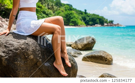Sunlit scene with a woman in white attire sitting on a large rock by a tropical beach, clear turquoise water, lush greenery, and blue sky creating a serene and relaxing atmosphere 125834177