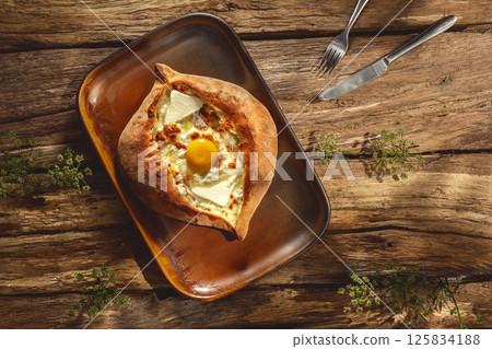 A traditional khachapuri rests on an authentic plate, basking in sunny light against a rustic wooden background, highlighting its golden crust and gooey cheese filling 125834188